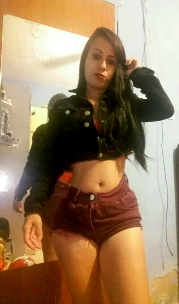 Young woman posing in a mirror selfie wearing a black cropped jacket and maroon shorts in a bedroom.